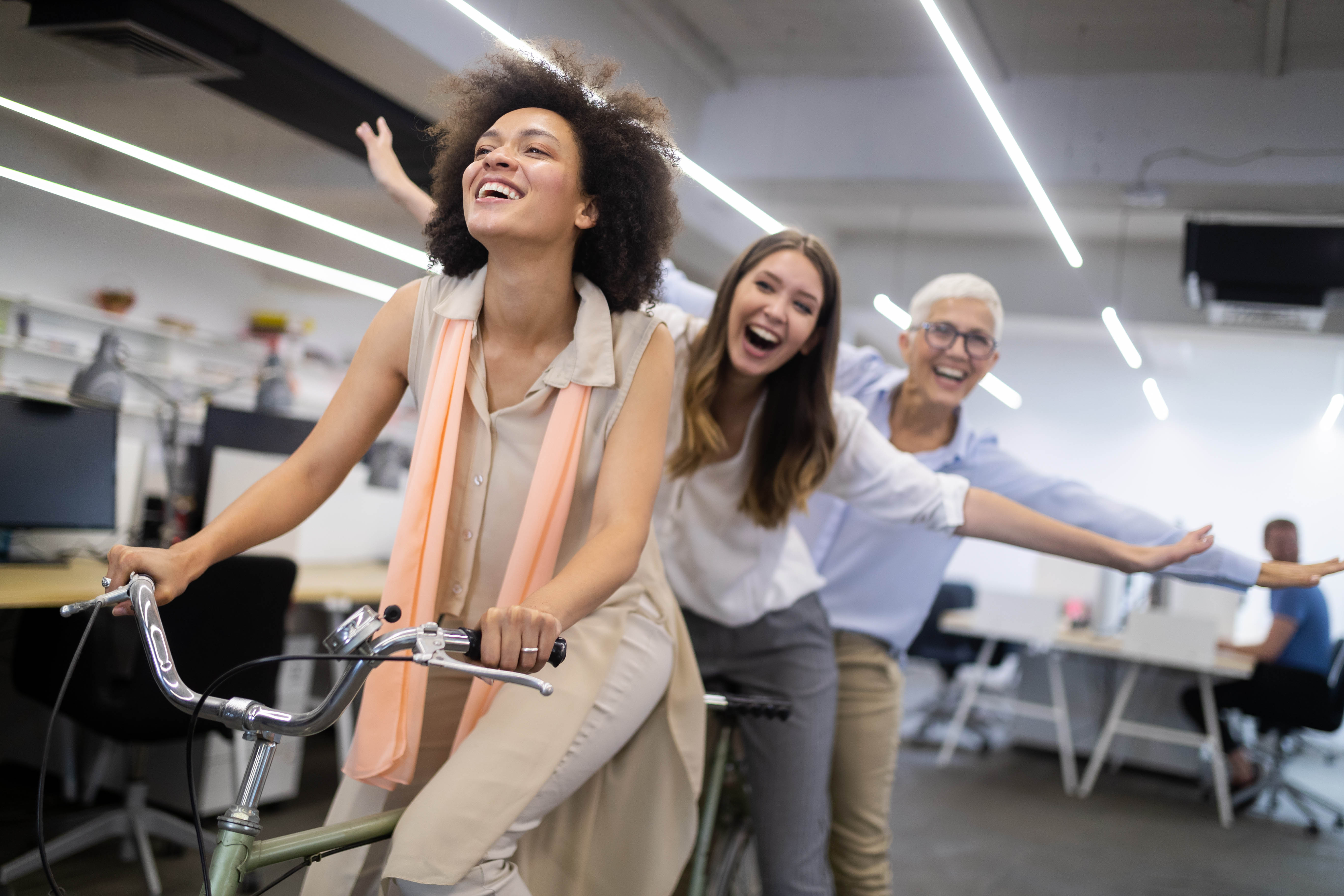 3 women of various ages riding a bike and laughing through the office, the two women on the back of the bike have their arms wide like they are flying 3 women of various ages riding a bike and laughing through the office, the two women on the back of the bike have their arms wide like they are flying